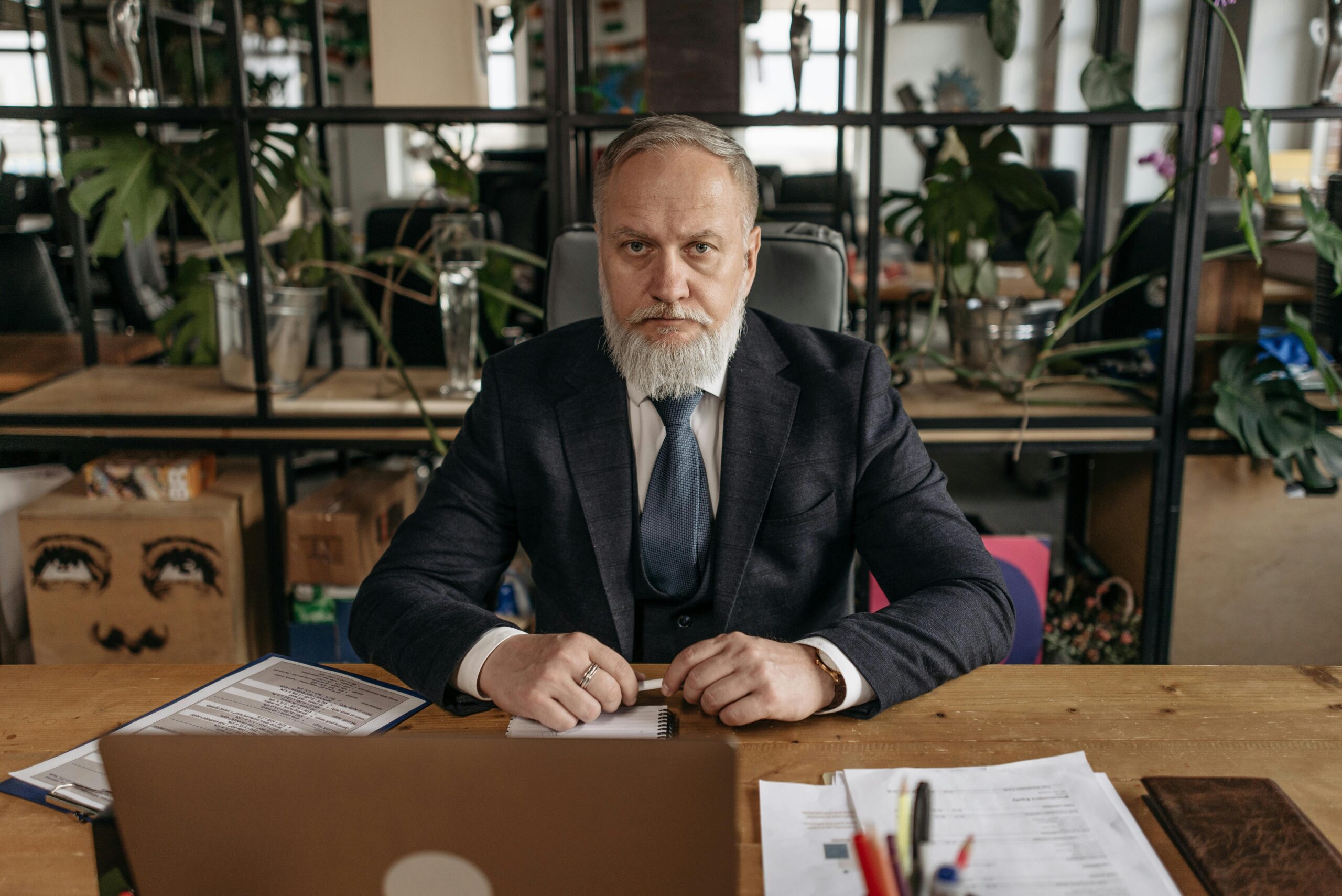 Elderly businessman in a suit at his office desk with papers and a laptop, serious expression.